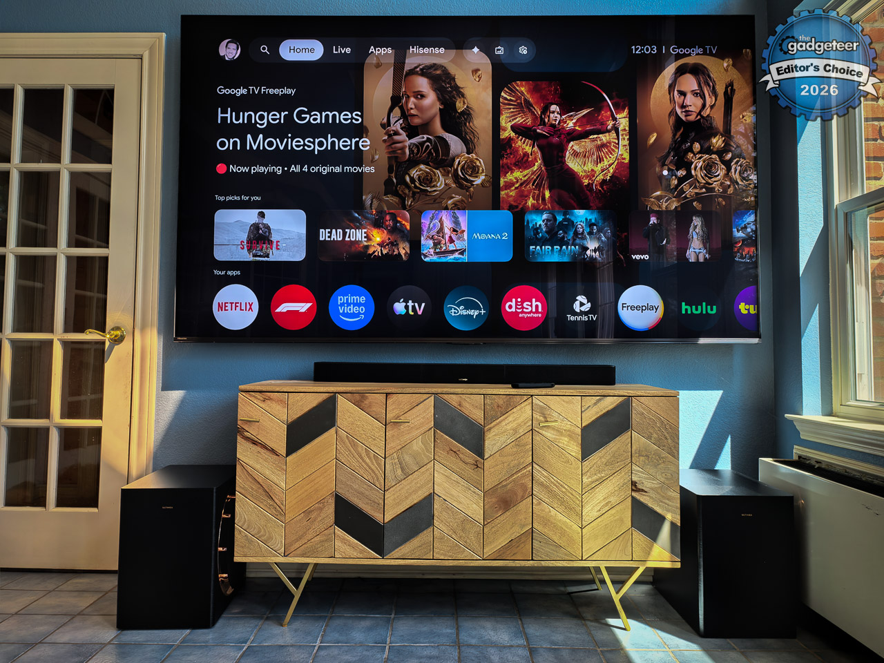 Ultimea SkyWave X100 Dual 9.2.6 wireless surround sound system fully set up in a living room, showing the 43.3-inch soundbar below a large-screen TV, four compact wireless surround speakers positioned around the seating area, and two 10-inch wireless subwoofers with black woodgrain finish and gold accents flanking the couch