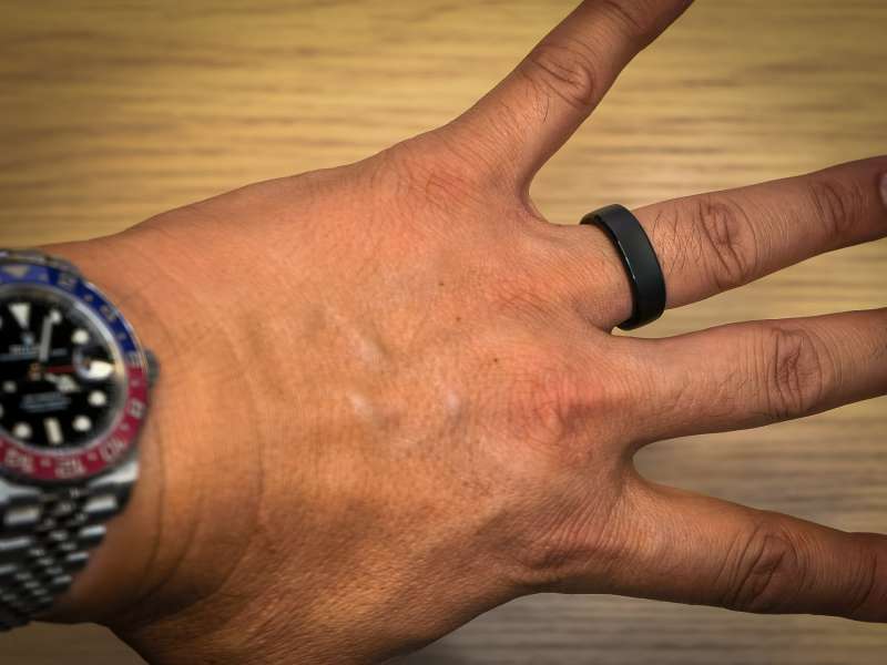 RingConn Gen 3 smart ring in black worn on the index finger of a man's outstretched hand, with a Pepsi-bezel GMT watch on the wrist, photographed on a wood desk