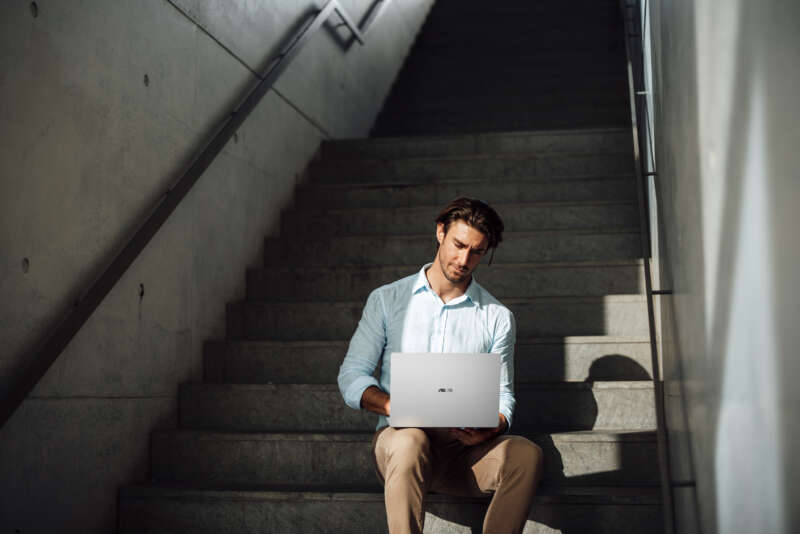 A man uses a silver ASUS ExpertBook A7 while sitting on concrete steps.