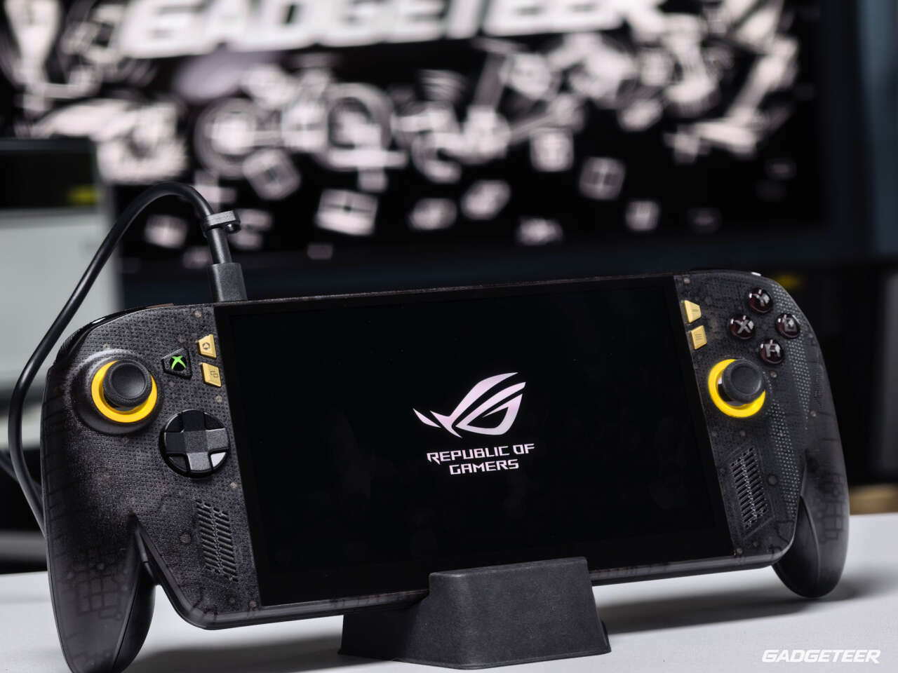 Black and gold ASUS ROG Xbox Ally X20 on a stand with its display powered on.