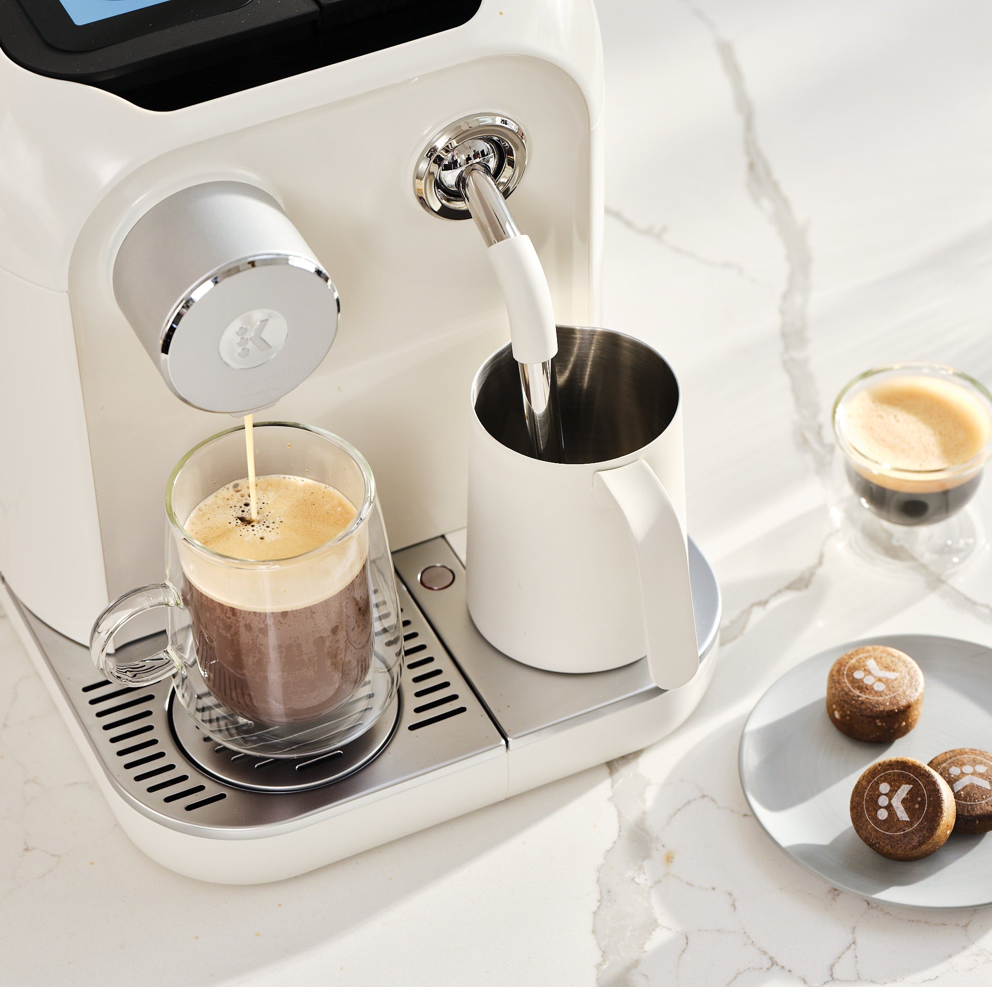 Keurig Alta brewing coffee beside its milk pitcher, with espresso and AltaRounds on the counter.