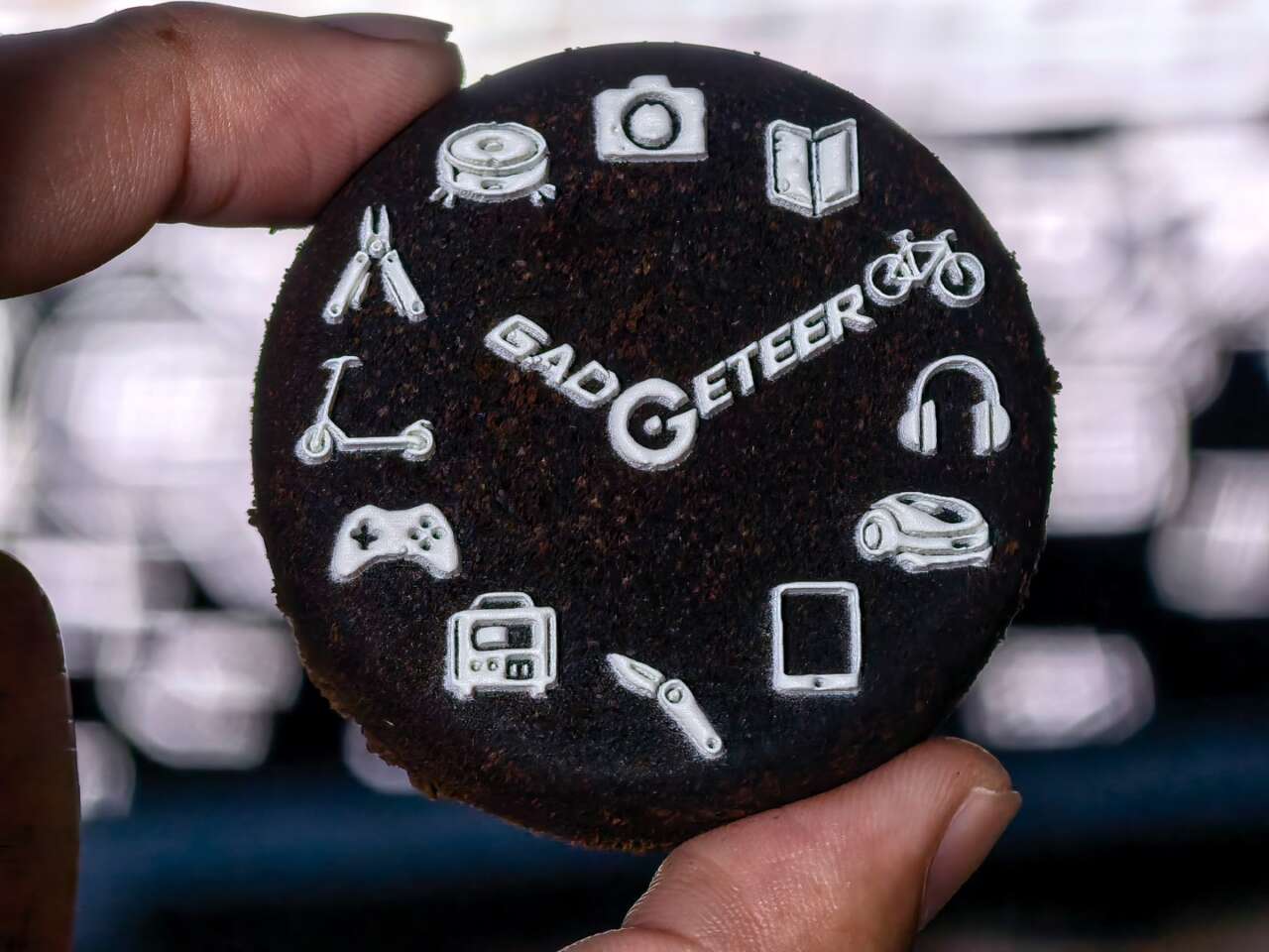 A used espresso coffee puck held between fingers, with The Gadgeteer logo and icons printed in white using the xTool O1 Omni.