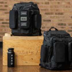 Two black KNKG APEX backpacks beside a wooden gym box