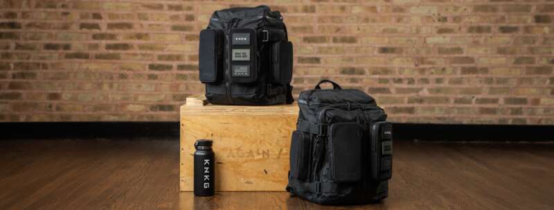 Two black KNKG APEX backpacks beside a wooden gym box