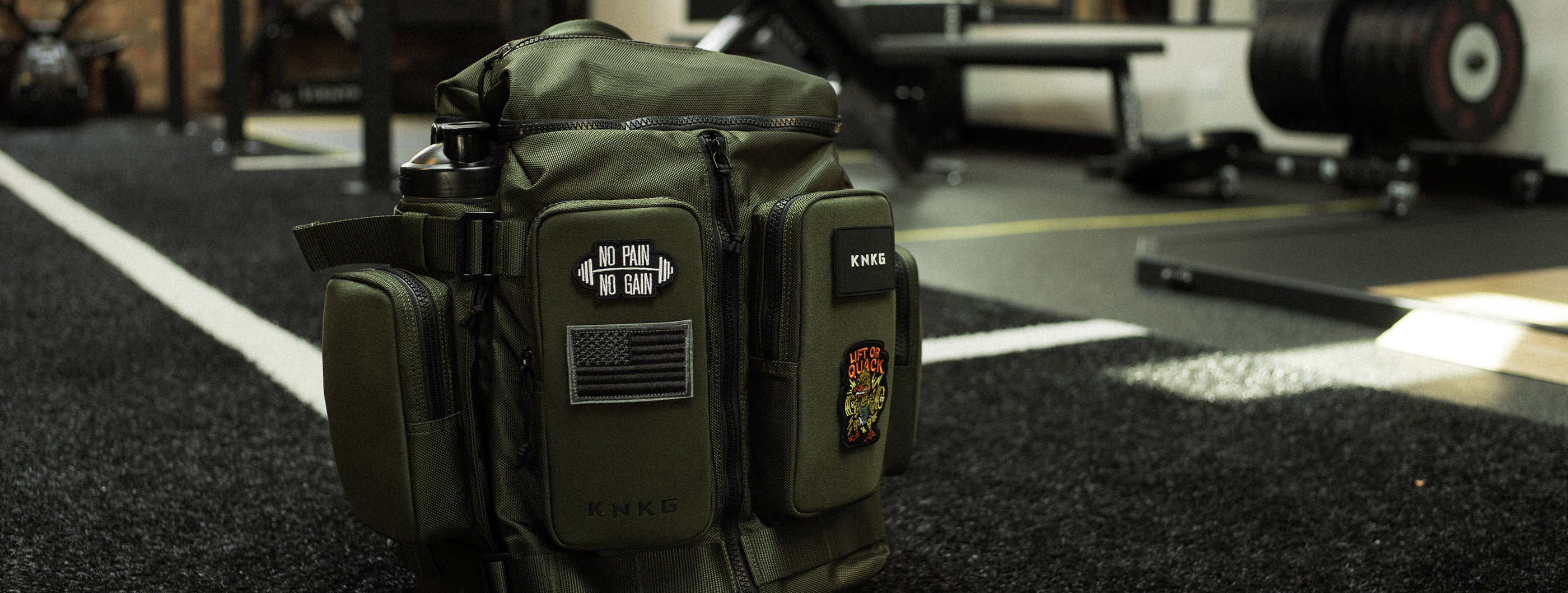 Olive KNKG APEX backpack with patches on its front panels and a bottle in a side pocket