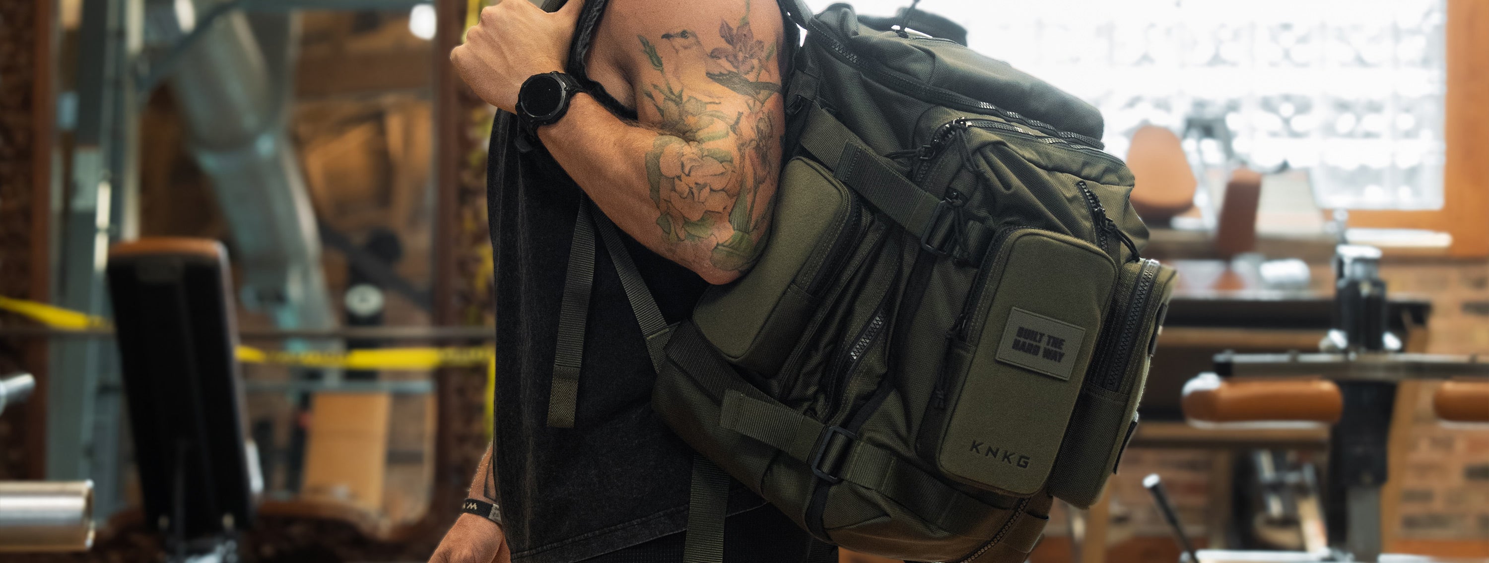 Person carrying an olive KNKG APEX backpack over one shoulder in a gym