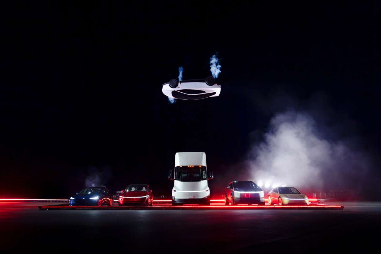 An inverted Tesla Roadster above a truck and car lineup during a night-time demonstration.