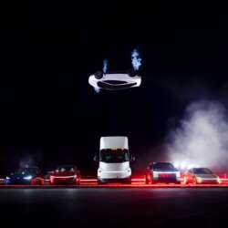 An inverted Tesla Roadster above a truck and car lineup during a night-time demonstration.