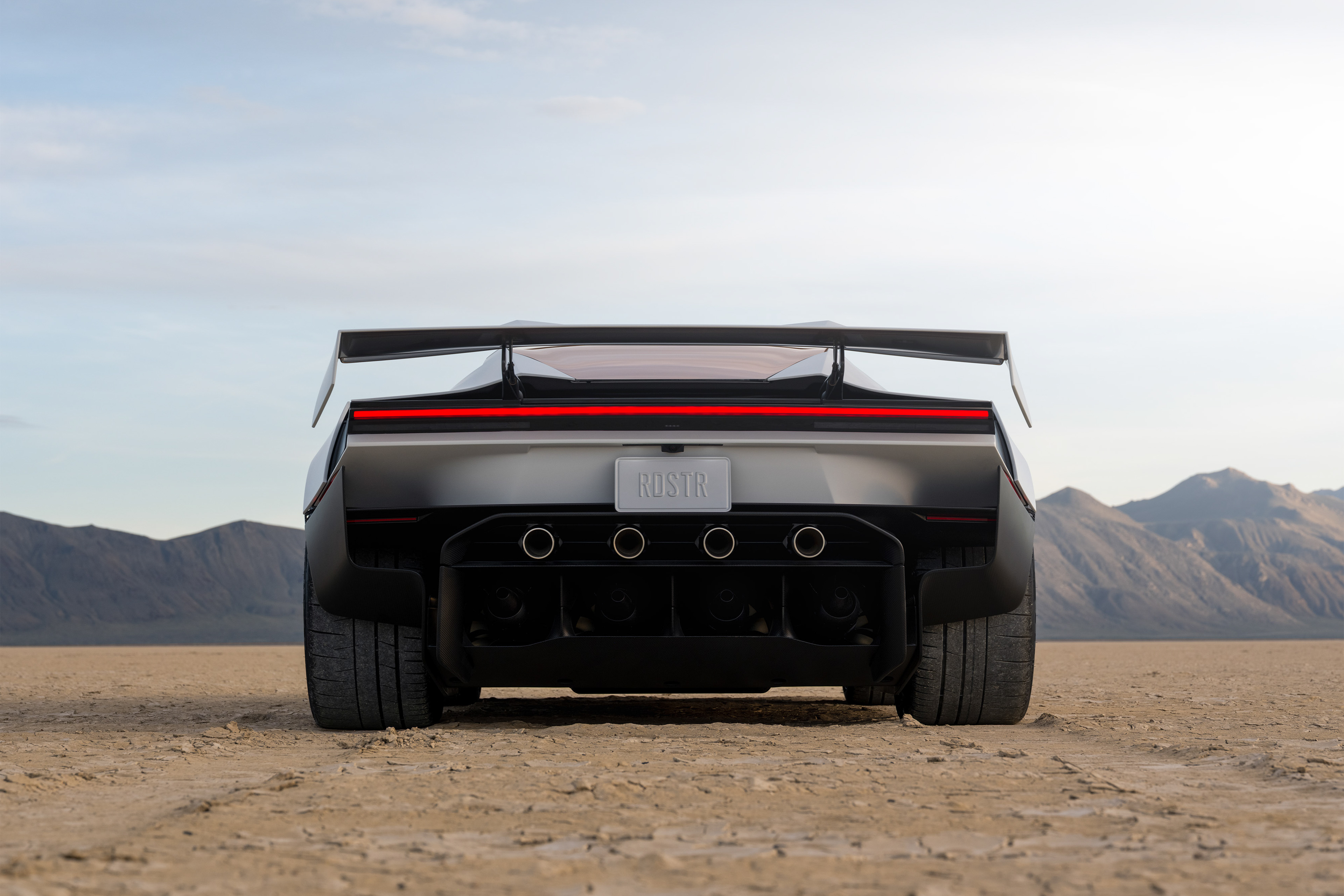 Rear of a silver Tesla Roadster with a large wing and red light bar on a dry lakebed